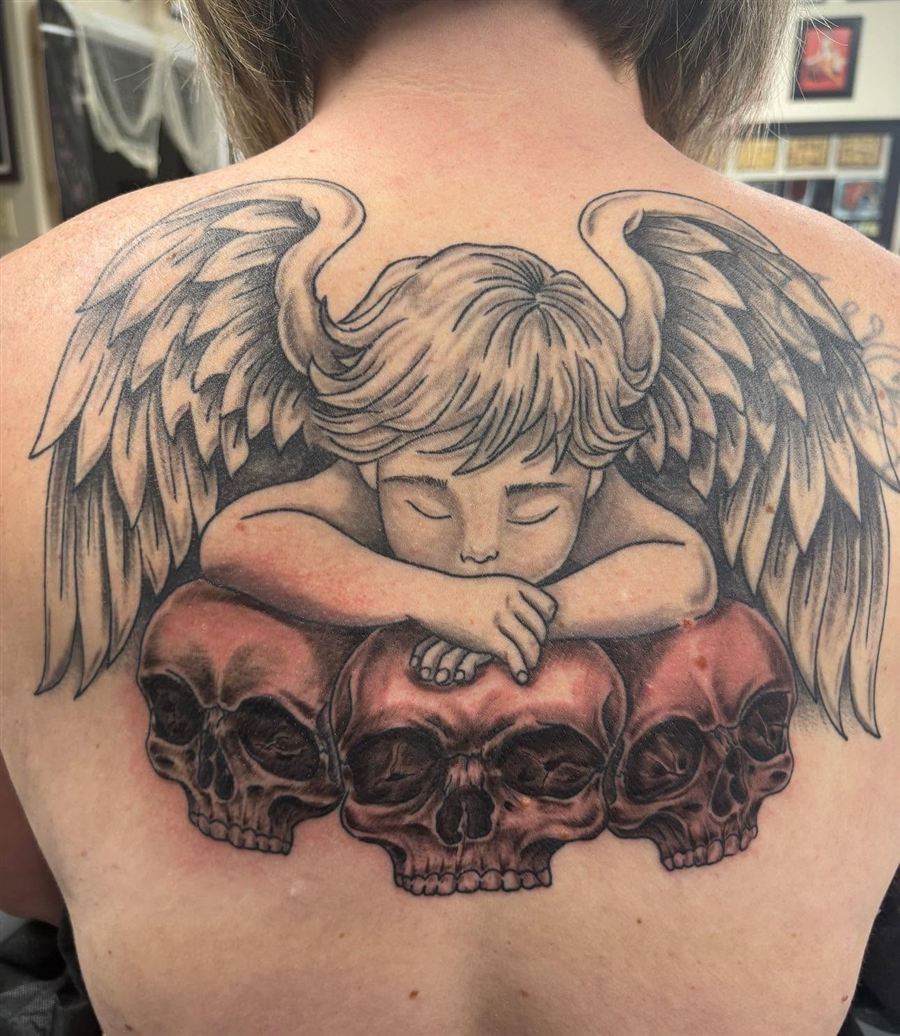 Black and grey cherub angel resting on skulls upper back tattoo
